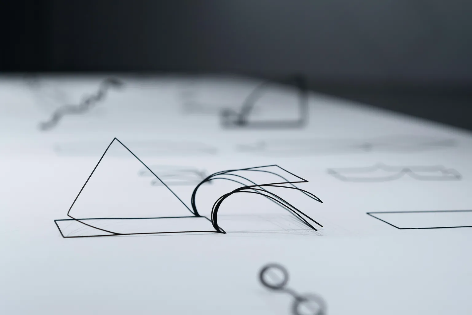 Des formes tracées en fil de fer noir, un triangle et des courbes, posées sur une table blanche.